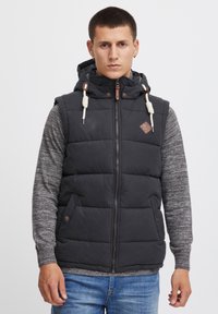 Chaleco de plumas negro con capucha, equipado con una cremallera frontal, bolsillos laterales y detalles de cuero marrón. Usado sobre una camiseta de manga larga gris.