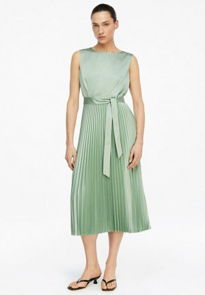Kobieta w bezrękawnikowej, jasnozielonej plisowanej sukience midi z paskiem w talii, stojąca boso na białym tle.