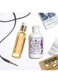 Aceite para peinar Bumble and Bumble en un frasco de vidrio transparente con detalles dorados, y Crema Definidora de Rizos en un frasco blanco con gráficos morados.