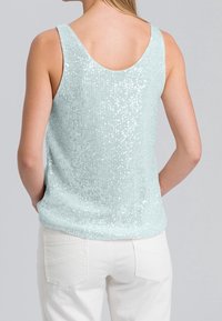Débardeur en sequins vert menthe avec un dos en scoop, présentant un tissu texturé et une coupe décontractée. Associé à un pantalon en denim blanc.