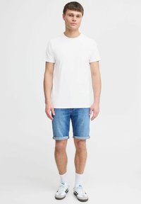 Ung mand står iført en ensfarvet hvid t-shirt, opskruede denim shorts, hvide sokker og hvide sneakers med sorte striber.