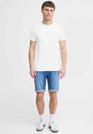 Bărbat tânăr stând, îmbrăcat cu un tricou alb simplu, pantaloni scurți din denim rulați, șosete albe și adidași albi cu dungi negre.