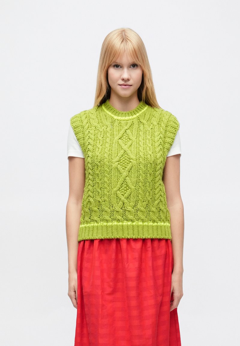 Laimiņzaļš adīts džemperis bez piedurknēm ar teksturētiem pinuma rakstiem, ribainu apakšmalu un īsām piedurknēm, komplektā ar plūstošu sarkanu svārku.