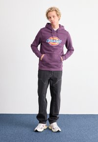 Violetinis džemperis su priekiu kišene ir grafikiniu logotipu. Derinamas su tamsiais džinsais, turinčiais kelis siūlus, ir baltais sportbačiais su violetiniais akcentais.