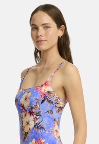 Un mannequin avec de longs cheveux bruns porte une robe florale bleu clair, posant avec confiance avec une expression neutre devant un fond blanc.