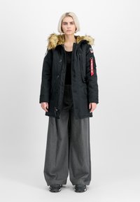 Črn parka s kapuco oblečeno z krznom, sprednjimi gumbi, več žepi in rdečo oznako. Kombinirana z širokimi sivimi hlačami in črnimi čevlji.
