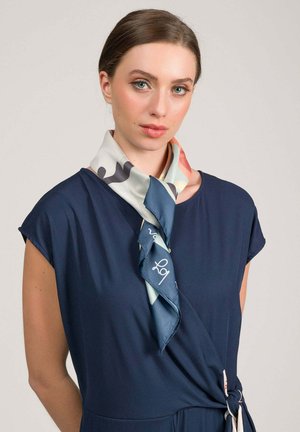 Donna con pelle chiara e capelli castani raccolti, che indossa un vestito blu navy e una sciarpa di seta multicolore annodata intorno al collo.