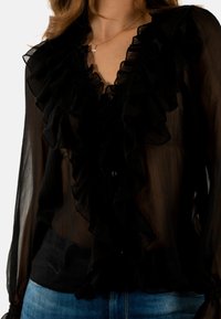 Blusa nera e trasparente con frontale arruffato, maniche lunghe e chiusura con bottoni. Presenta un tessuto testurizzato e un design a strati. Abbinata a jeans blu.