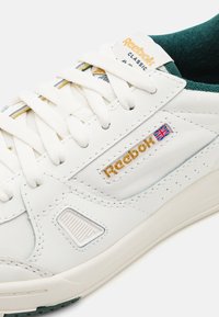 Balti odiniai Reebok Classic sportiniai bateliai su baltais raišteliais, auksiniu Reebok užrašu ir maža Jungtinės Karalystės vėliava šone.