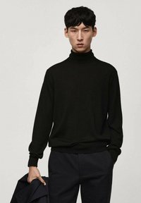 Svart turtleneck-tröja gjord av mjukt, stickat tyg. Har långa ärmar och en avslappnad passform, tillsammans med mörka byxor och håller en jacka.