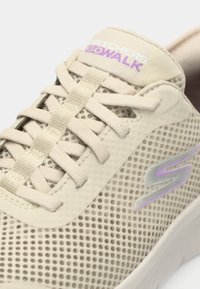 Μπεζ αθλητικό παπούτσι Skechers GOWALK με υλικό από δίχτυ, επίπεδα κορδόνια, επενδυμένη γλώσσα και διακριτικό μοβ λογότυπο στο πλάι.