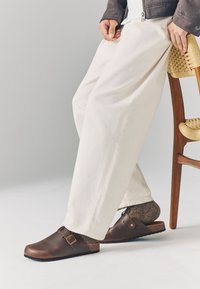 Braune Leder-Clogs mit Schnallendetail, Korkfußbett und strukturierten braunen Socken, getragen zu locker sitzenden beigen Hosen neben einem Holzstuhl.