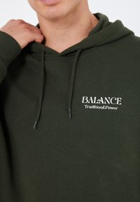 Felpa verde con cappuccio, polsini e orlo a costine, presenta la scritta bianca "BALANCE Tradition & Power" sul petto. Tessuto con texture morbida.