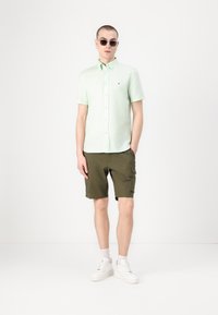 Világos zöld, rövid ujjú ing, khaki cargo rövidnadrág és fehér sportcipő. A modellen kerek napszemüveg van. Egyszerű dizájn, laza szabás.