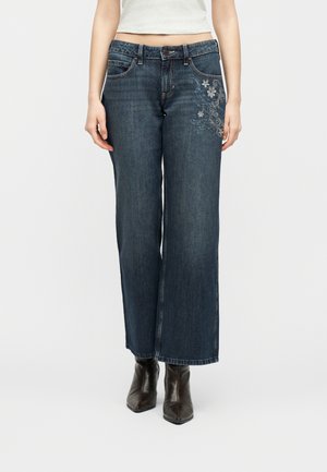 Kvinde iført mørkeblå jeans med vide ben og blomsterbroderi på højre lår samt sorte spidse ankelstøvler.