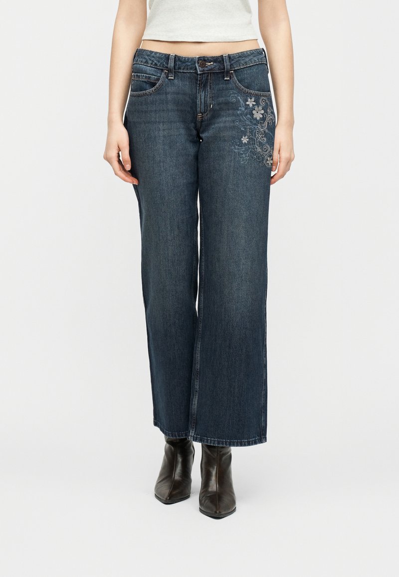 Γυναίκα που φοράει σκούρο μπλε τζιν με φαρδιά μπατζάκια και κεντήματα με λουλούδια στο δεξί μηρό, καθώς και μαύρα μυτερά μποτάκια αστραγάλου.