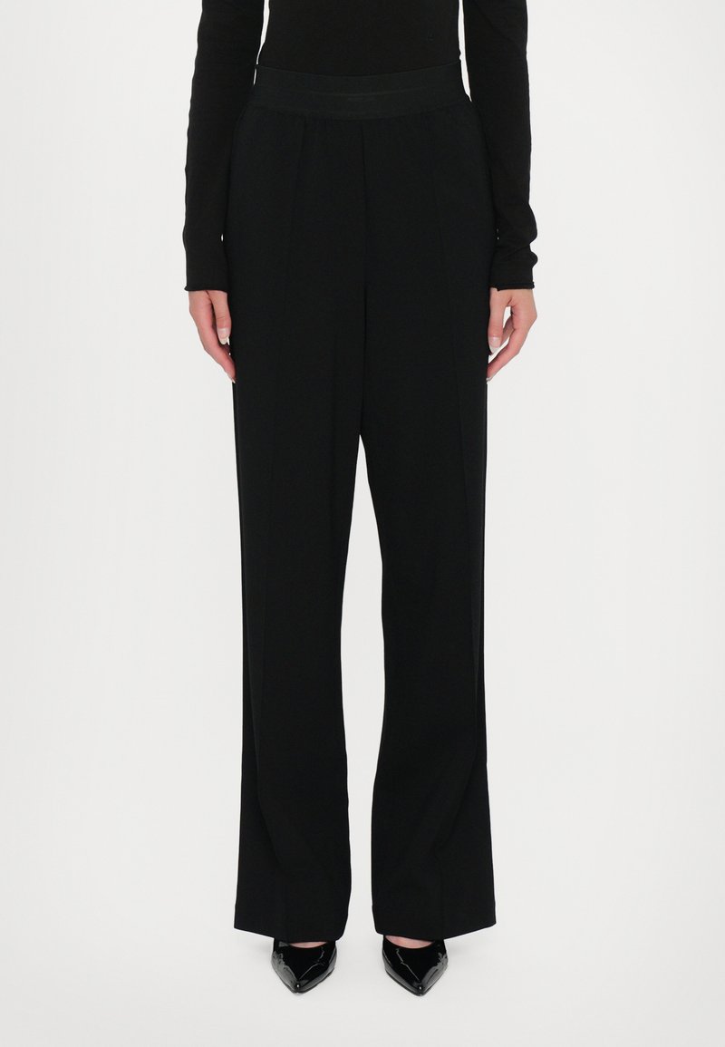 Pantaloni neri a gamba larga con una texture liscia e una vita alta. Presentano pinces frontali e sono abbinati a scarpe nere a punta.