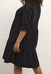 Schwarzes knielanges Kleid mit einem gestuften Rock, 3/4-Ärmeln mit Knöpfen, aus leichter, weicher und fließender Stoffqualität.