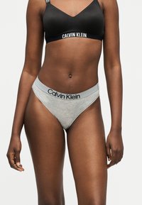 Γυναίκα που φοράει μαύρο bralette Calvin Klein και γκρι εσώρουχο Calvin Klein, στέκεται μπροστά σε ένα απλό ανοιχτό φόντο.