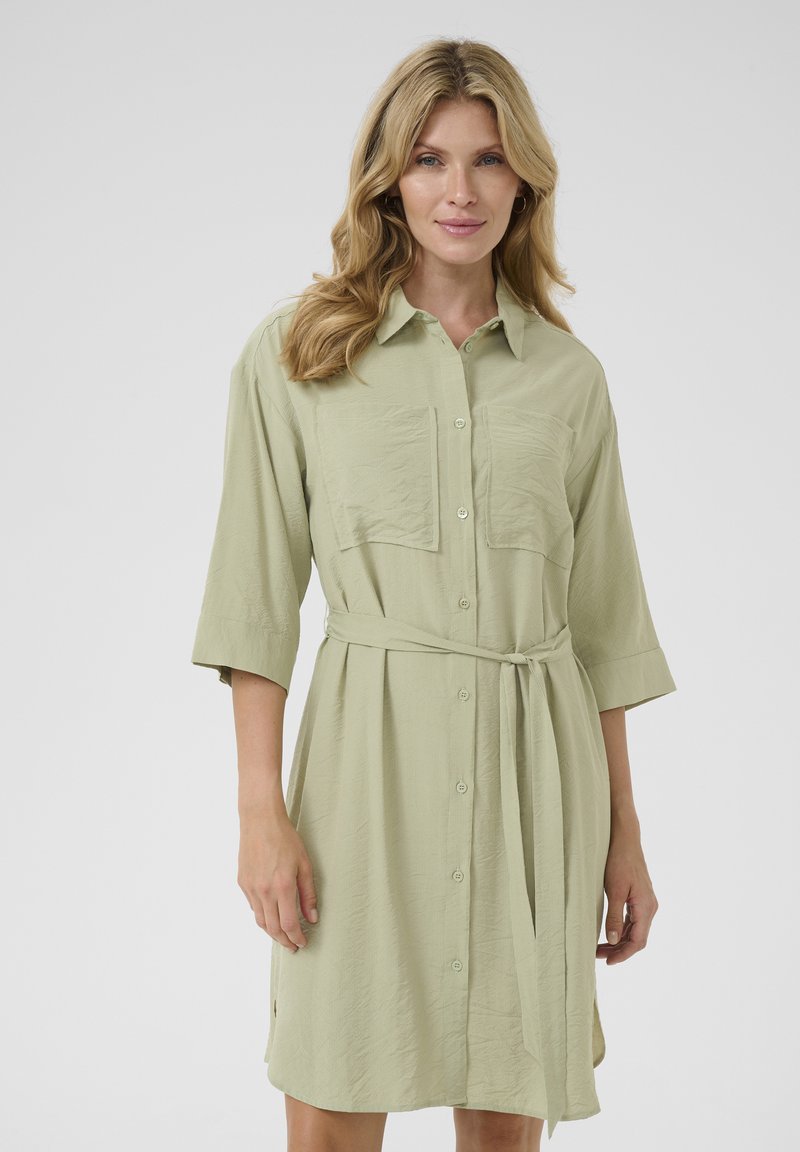 Abito camicia verde chiaro con bottoni sul davanti, due tasche sul petto e vita legata. Presenta maniche 3/4 e un tessuto morbido e testurizzato.