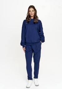 Granatowa bawełniana bluza z kapturem o luźnym kroju, w zestawie z dopasowanymi dresami z plisami; noszona z białymi sneakersami.