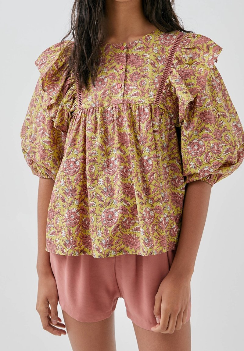 Blouse à motifs floraux jaune et rose, avec manches volantes, col à boutons et coupe décontractée. Associée à un short rose en dessous.