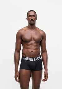 Črne bokserice s belim logotipom "CALVIN KLEIN" na pasu, narejene iz gladkega materiala, prilegajoč dizajn, nosi jih moški brez majice.