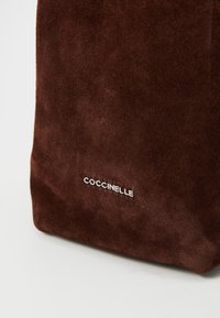 Ruda zomšinė rankinė su minkštu paviršiumi, turinti sidabrinį "Coccinelle" logotipą, stačiakampio formos ir siuvinėtomis kraštinėmis.