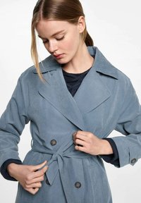 Svetlo modrý zamakový trenčkot s veľkým golierom, dvojitým gombíkovým zapínaním a opaskom v páse. Obsahuje dlhé rukávy s tmavými pletenými manžetami.