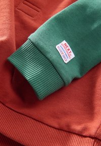 Grüner, gerippter Bund an einem orangefarbenen Kleidungsstück, mit einem Etikett, das "NEXT" und Produktdetails zeigt. Strukturiertes Gewebe in gefalteten Schichten dargestellt.