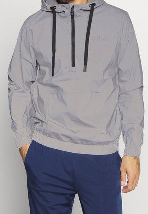 Mann trägt eine hellgraue FILA-Windjacke mit Kapuze und schwarzen Kordelzügen sowie marineblaue Hose, Hände in den Taschen, vor einfachem Hintergrund.