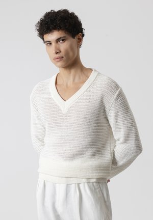 Persona joven con cabello rizado que lleva un suéter de punto blanco holgado con cuello en V y pantalones blancos, de pie con las manos detrás de la espalda sobre un fondo gris.