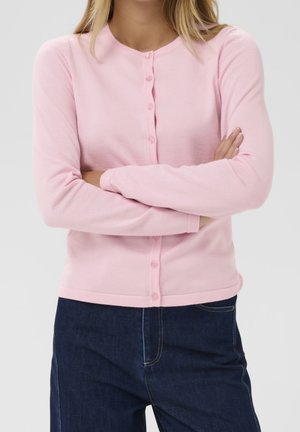 Person iført en lyserød cardigan med knapper og lange ærmer samt mørkeblå jeans, krydsede arme ved livet.