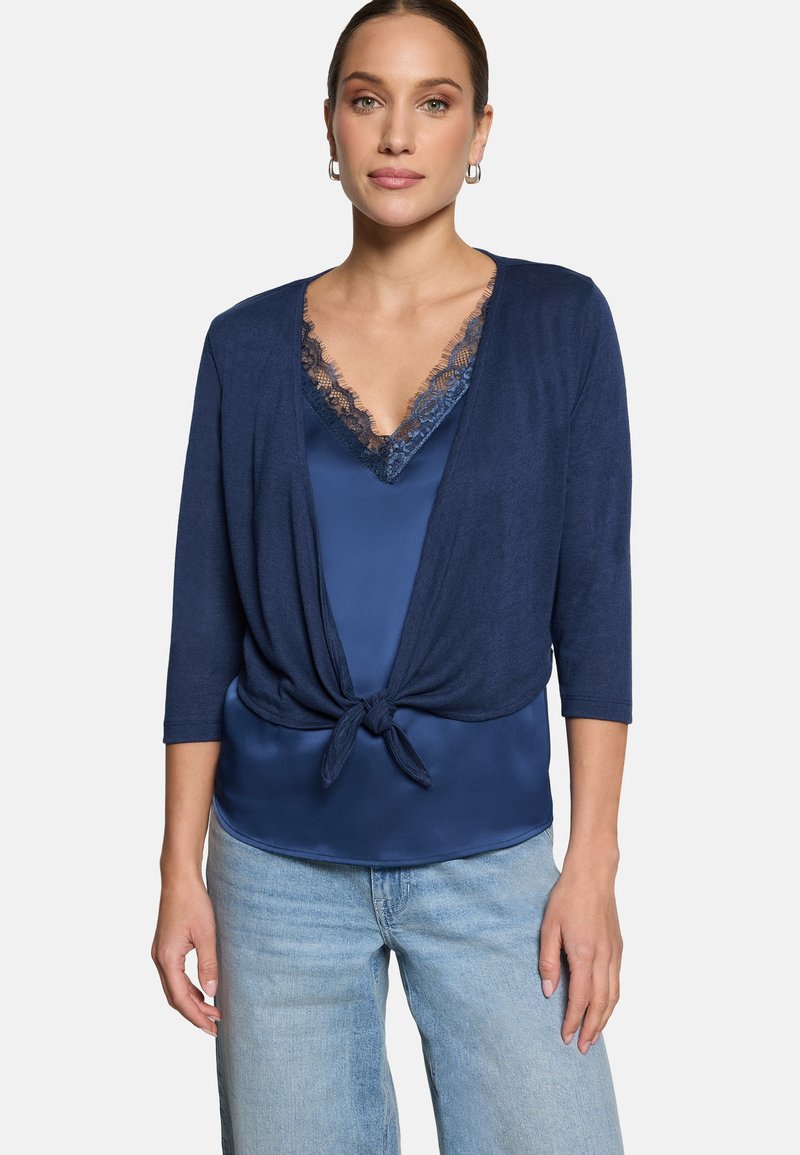 Donna che indossa un top di satin blu navy con rifiniture in pizzo, un cardigan blu navy annodato in vita e jeans azzurri, in piedi contro uno sfondo bianco.