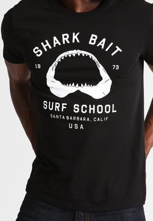 Schwarzes Baumwoll-T-Shirt mit weißem Motiv eines Haifischmauls und dem Text "SHARK BAIT SURF SCHOOL" sowie fettgedruckten Ortsangaben.