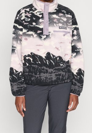 Person iført en Columbia fleecepullover i sort, hvid og pink mønster med trykknapper og en lynlåslomme på brystet.