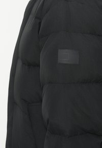 Sort pufferjakke med en quiltet design. Har et struktureret stof og et rektangulært logo-patch påærmet.
