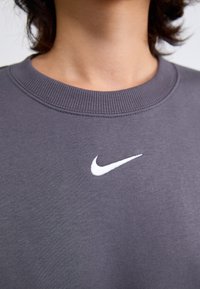 Grijze sweatshirt van zacht materiaal, met een wit Nike-logo op de borst en een geribbelde neklijn. Soepele textuur met minimale details.