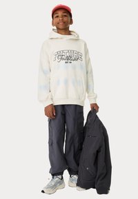 Sweat à capuche crème avec des accents tie-dye bleus et un graphique "FUTURE Trailblazer", associé à un pantalon cargo noir et des baskets blanches. Tenant une veste noire.