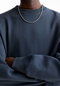 Felpa blu scuro con colletto e polsini a costine, caratterizzata da una piccola stampa del logo sulla manica. Abbinata a una collana in argento.