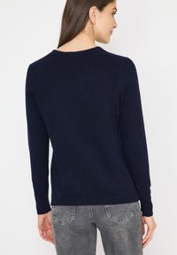 Marineblaues Strickpullover mit langen Ärmeln, Rundhalsausschnitt und gerippten Bündchen. Weiche Textur und figurbetonter Schnitt, über grauen Jeans getragen.