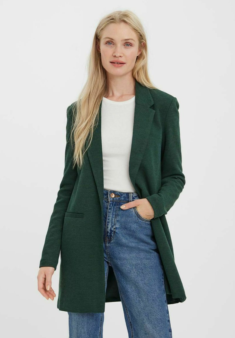 Grüne Blazer aus weichem Material, mit Reverskragen, einer einzelnen Fronttasche und offenem Design, getragen über einem weißen Oberteil und blauen Jeans.