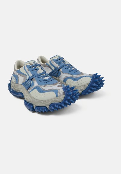 Par de zapatos deportivos grises y azules con parte superior de malla y suelas azules gruesas con picos, diseñadas para un agarre en terrenos accidentados.