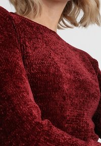 Burgundy-tyylinen kangas, jossa on pehmeä, samettimainen pinta. Ominaisuuksiin kuuluu pitkät hihat ja pyöreä kaula-aukko, joka esittelee rikasta väriä ja pehmeää pintaa.