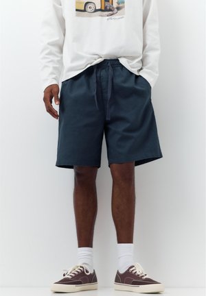 Muž nosiaci tmavomodré šortky so sťahovacou šnúrkou, biele tričko s dlhým rukávom s grafikou, biele ponožky a hnedé nízke tenisky.