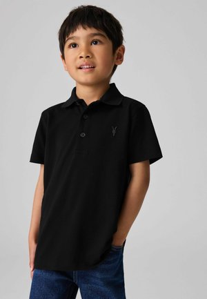 Polo noir en coton avec une patte de boutonnage à trois boutons, manches courtes et un design brodé sur la poitrine. Un jean en denim est visible.