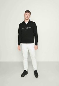 Svart genser med kvart glidelås, utstyrt med hvit og rød "TOMMY HILFIGER" logo. Brukt sammen med hvite bukser og svarte joggesko på en nøytral bakgrunn.