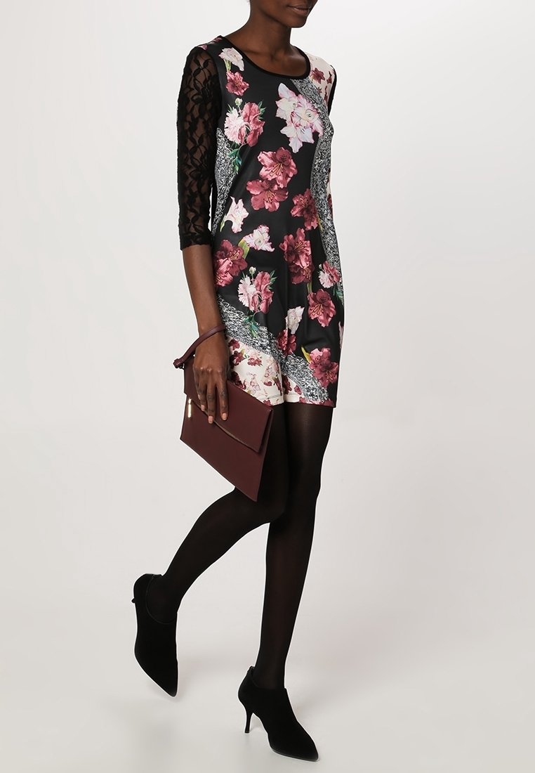 Femme vêtue d'une robe noire à fleurs avec des manches en dentelle, des collants noirs, des talons noirs, tenant une pochette bordeaux, marchant sur un fond uni.