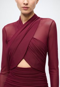 Robe portefeuille bordeaux avec manches longues transparentes, présentant un corsage à motifs croisés et une découpe au-dessus de la taille, fabriquée en tissu lisse.