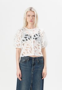 Chemisier blanc transparent avec des motifs de dentelle complexes, manches courtes bouffantes et encolure ronde, superposé sur un haut noir, associé à une jupe en jean.